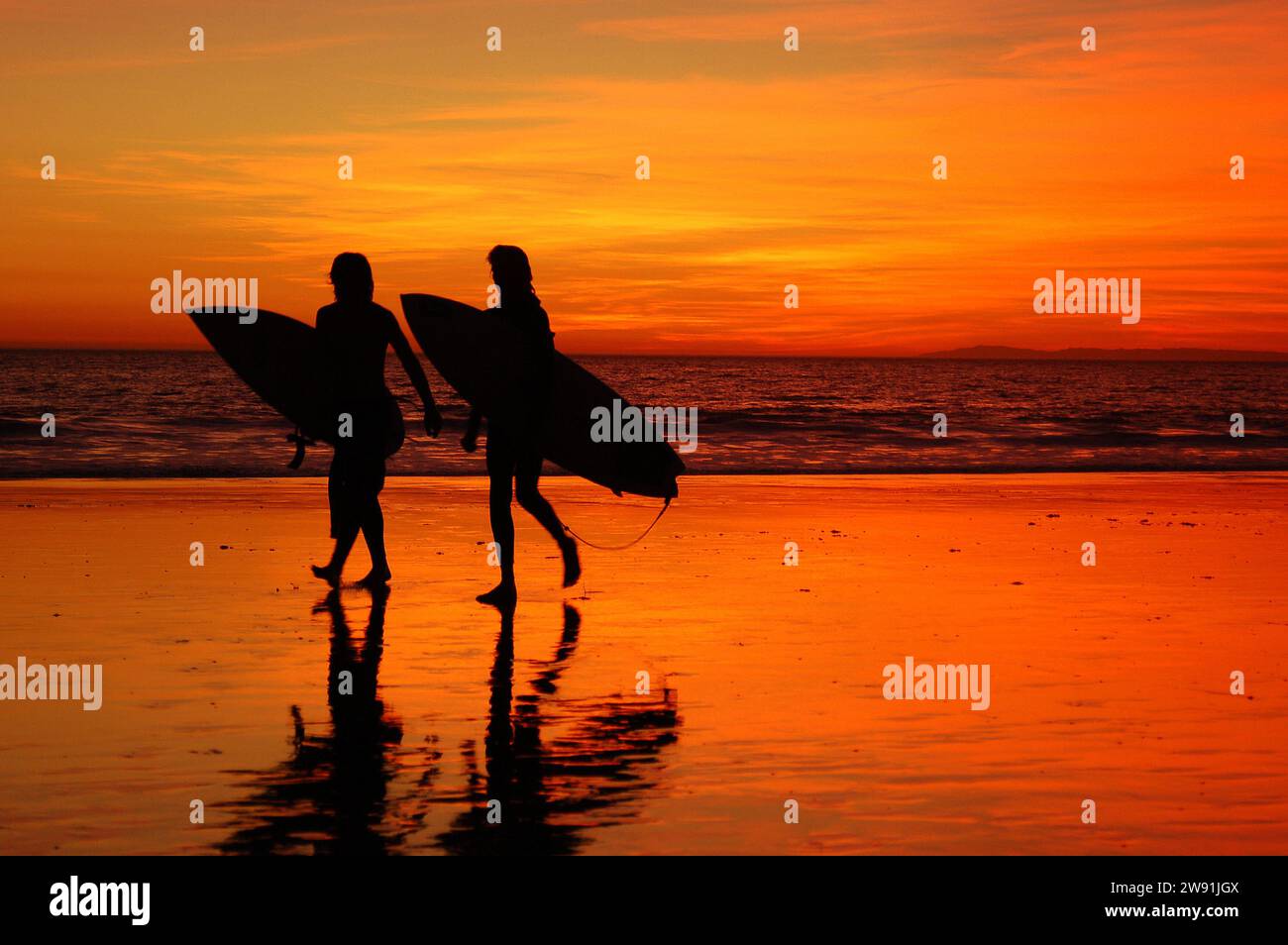 Due surfisti adolescenti, in silhouette dal cielo del tramonto, lasciano l'oceano portando le loro tavole da surf dopo una giornata di surf sulla spiaggia in California Foto Stock