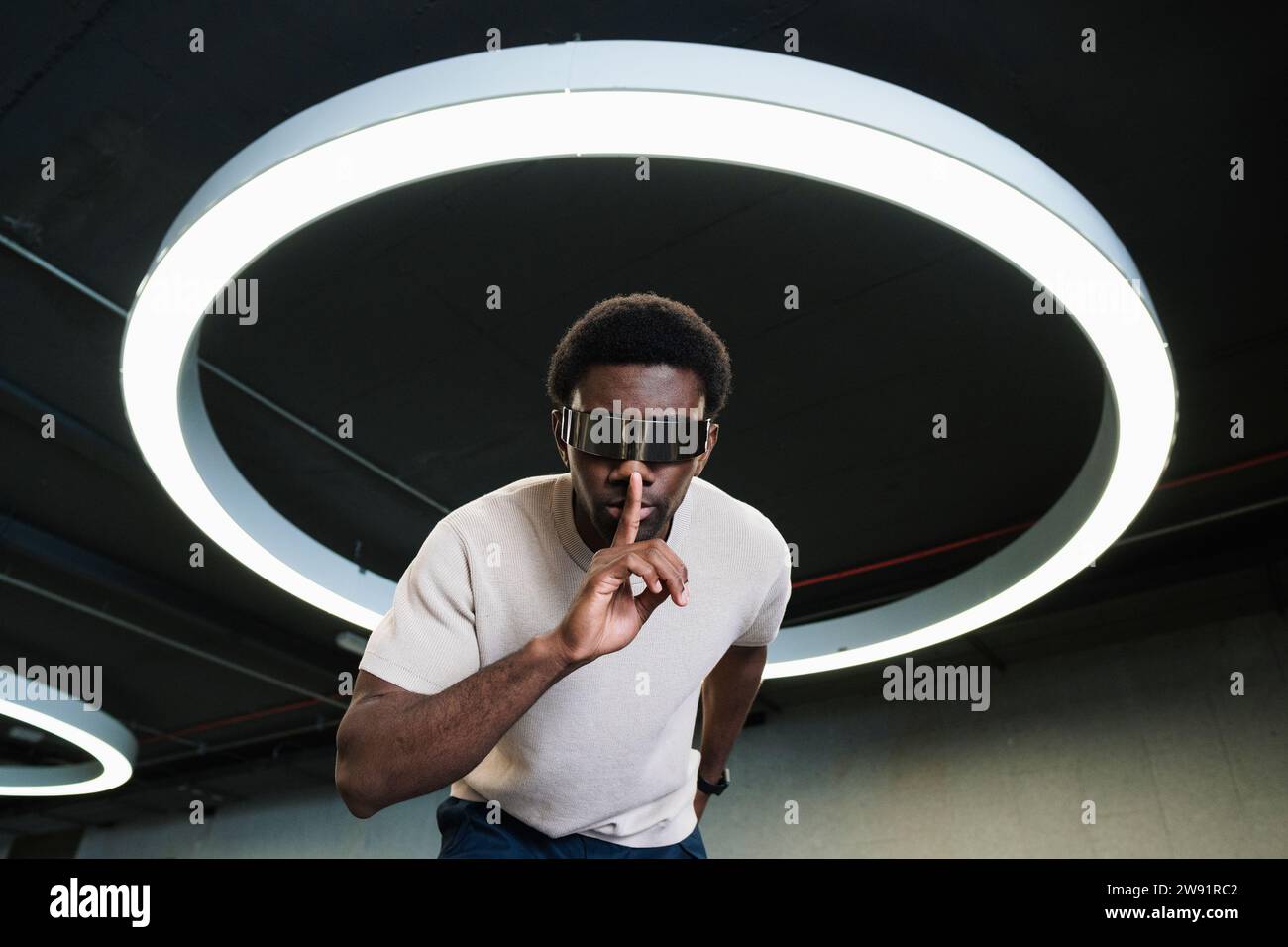 Giovane con futuristici bicchieri cyber sotto una moderna lampada ad anello che mette l'indice sulle labbra Foto Stock