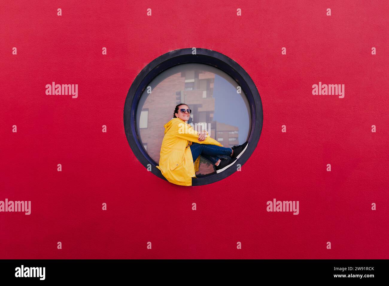 Donna con impermeabile giallo seduta in oblò su parete rossa Foto Stock