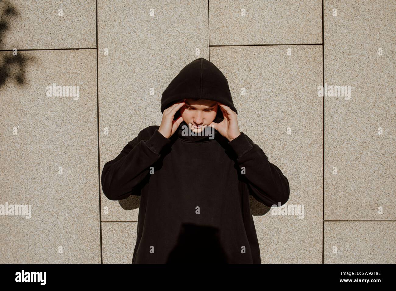 Ragazzo adolescente stressato che indossa una camicia nera con cappuccio davanti al muro Foto Stock