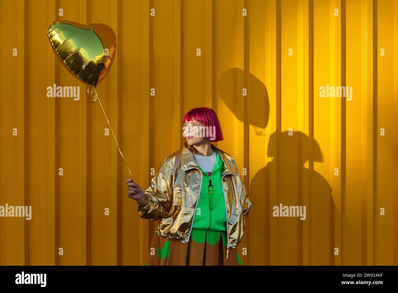 Donna anziana sorridente che tiene un palloncino a forma di cuore davanti alla parete gialla Foto Stock