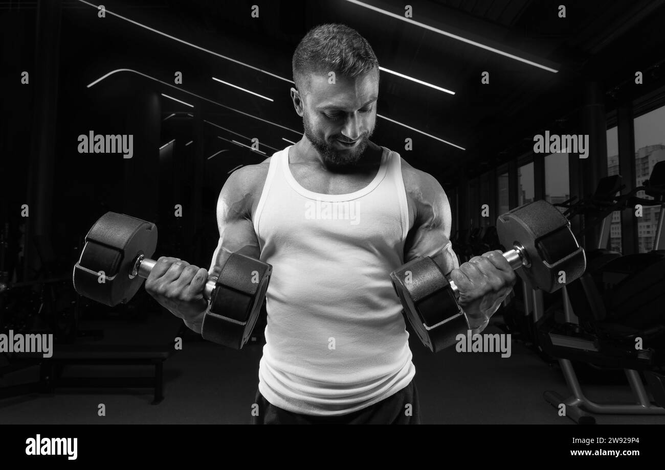 Uomo muscoloso con una t-shirt bianca lavora in palestra con manubri. Pompaggio bicipite. Concetto di fitness e bodybuilding. Supporti misti Foto Stock