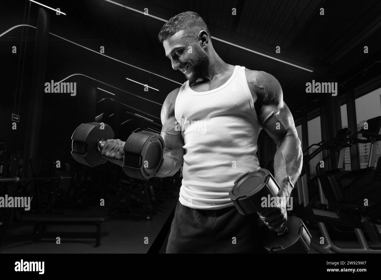 Uomo muscoloso con una t-shirt bianca lavora in palestra con manubri. Pompaggio bicipite. Concetto di fitness e bodybuilding. Supporti misti Foto Stock