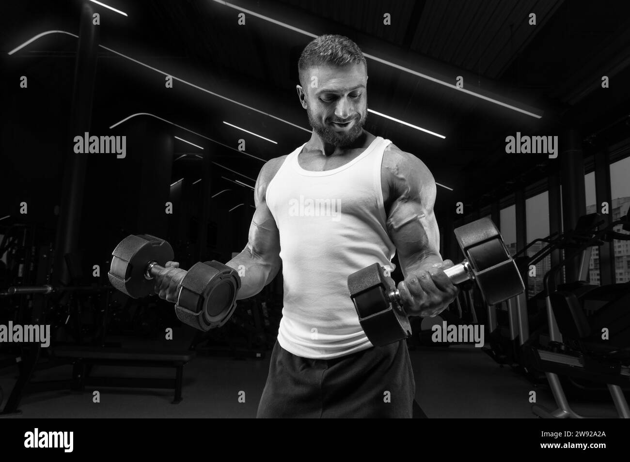 Uomo muscoloso con una t-shirt bianca lavora in palestra con manubri. Pompaggio bicipite. Concetto di fitness e bodybuilding. Supporti misti Foto Stock
