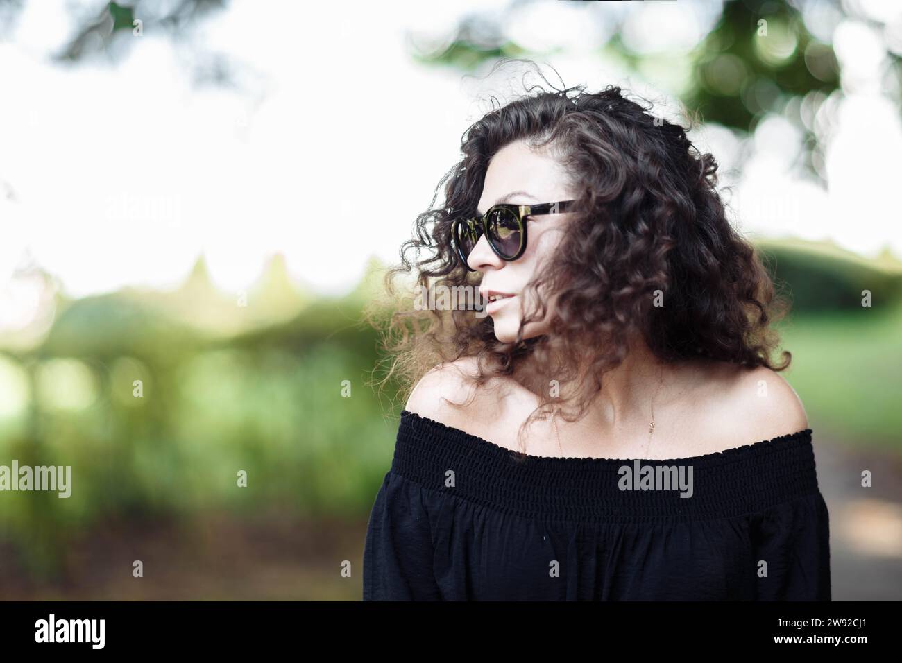 Una giovane donna riccia snella con un vestito nero e occhiali da sole si pone in una giornata di sole in un parco cittadino Foto Stock