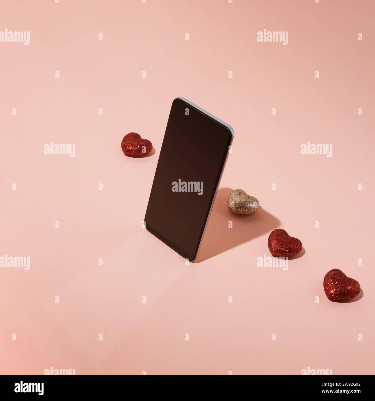 Schermo dello smartphone in piedi di fronte a una fila di cuori rossi scintillanti su sfondo rosa pastello. Concetto di app mobile e incontri online. Foto Stock