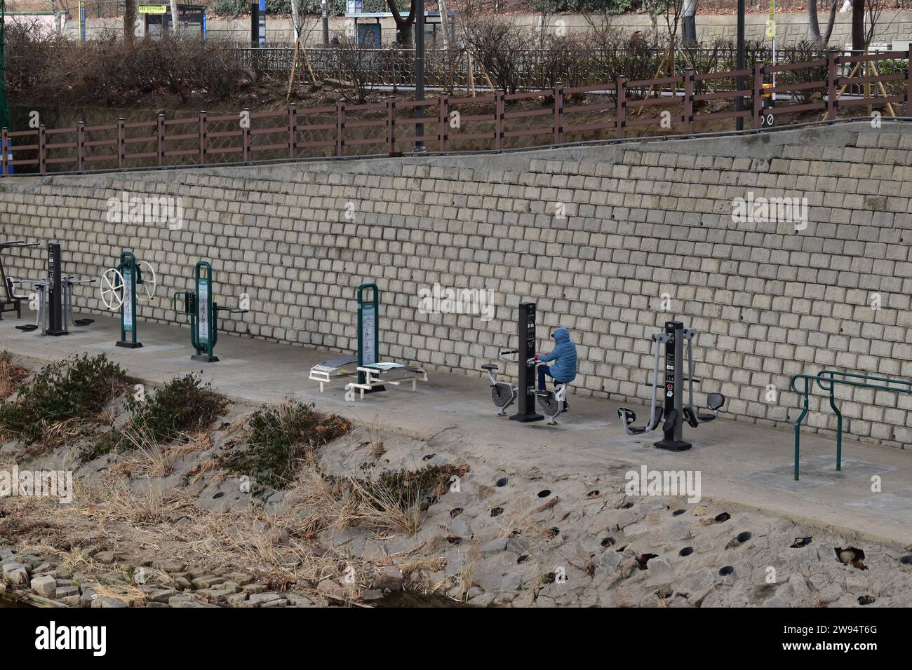 Persona in giacca blu che si esercita sulle attrezzature della palestra pubblica sulla riva di cemento di un piccolo canale nel quartiere di Seodaemun-GU Foto Stock