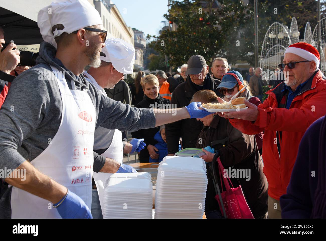 Pola, Croazia. 24 dicembre 2023. Persone che ricevono una porzione di un tradizionale risotto di pesce (risotto ai Fruitti di Mare) durante una tradizionale vigilia di Natale - materiale pubblicitario natalizio presso il mercato cittadino di Pola, Croazia, la vigilia di Natale del 24 dicembre 2023. Foto: SASA Miljevic/PIXSELL credito: Pixsell/Alamy Live News Foto Stock