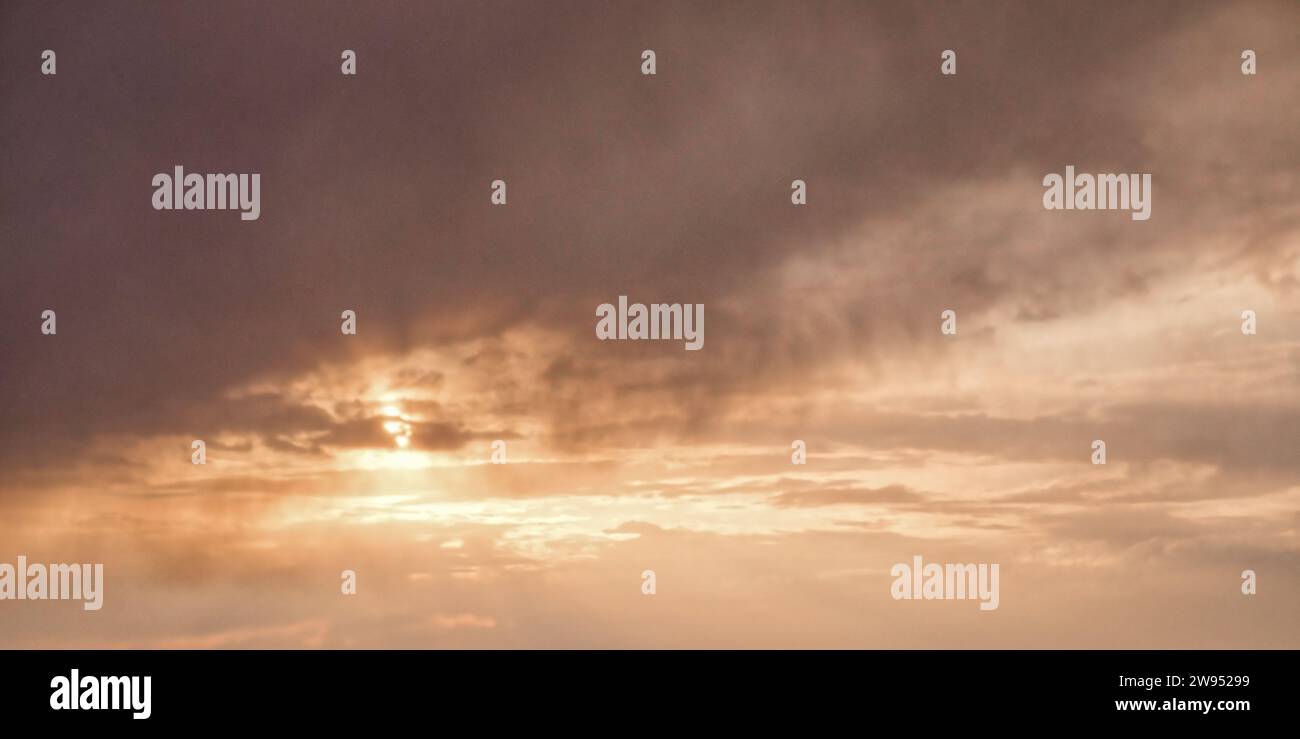 L'immagine mostra un cielo nuvoloso con il sole che sbircia attraverso, proiettando un caldo bagliore tra le nuvole scure. Foto Stock