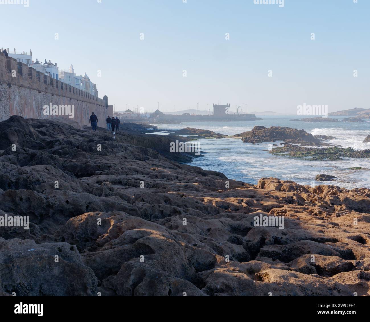 La gente cammina lungo la costa rocciosa con le mura della città della Medina e il forte più avanti, a Essaouira, in Marocco. 23 dicembre 2023 Foto Stock