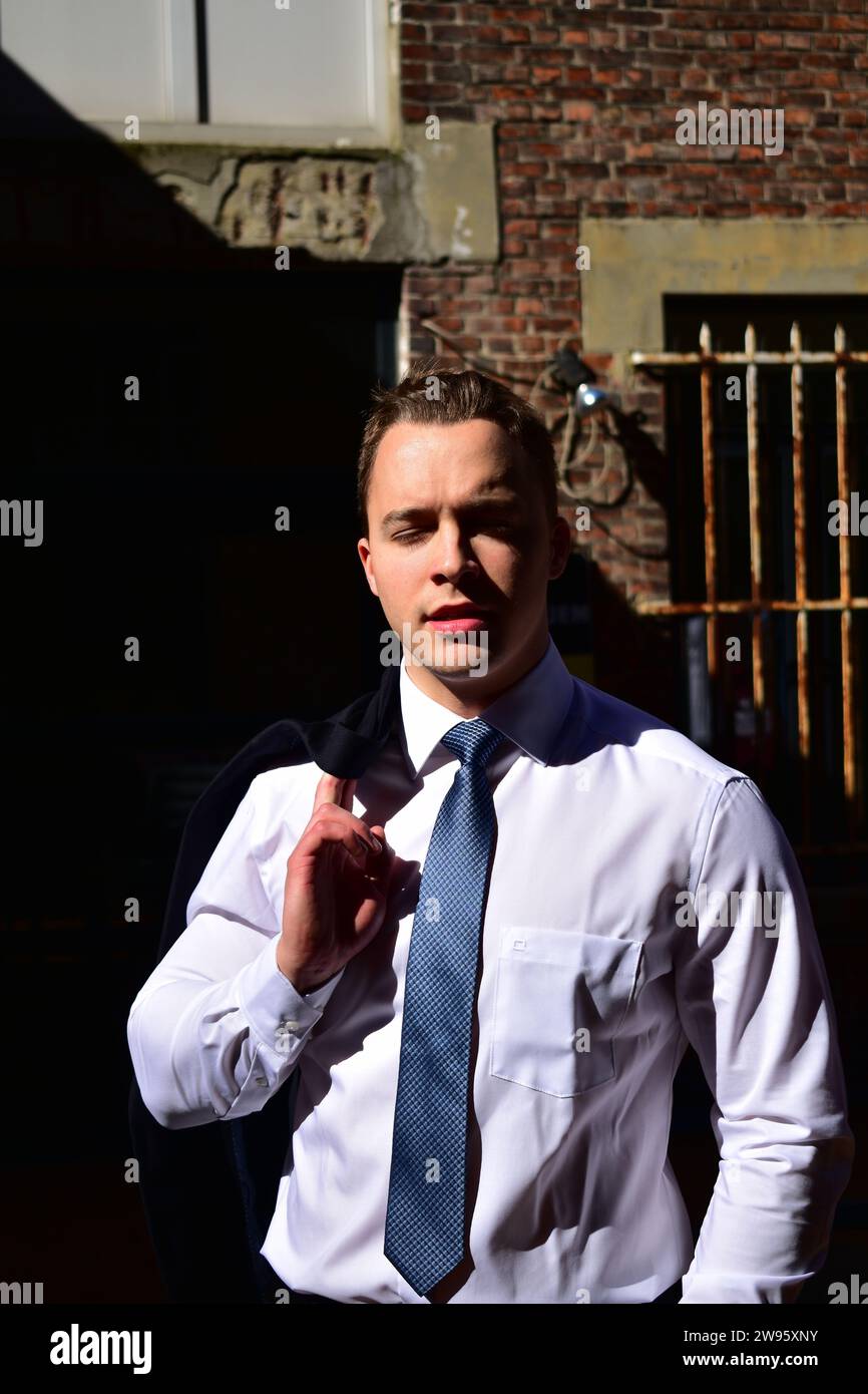 Giovane uomo bianco muscoloso che porta la giacca nera sulla spalla e indossa una camicia bianca con colletto e cravatta blu davanti a una parete di mattoni Foto Stock
