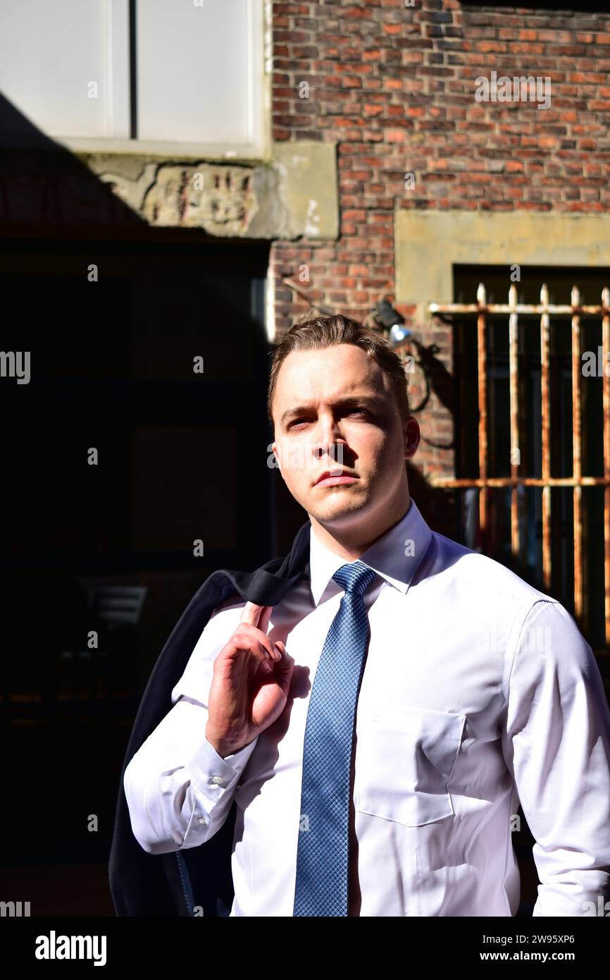 Giovane uomo bianco muscoloso che porta la giacca nera sulla spalla e indossa una camicia bianca con colletto e cravatta blu davanti a una parete di mattoni Foto Stock