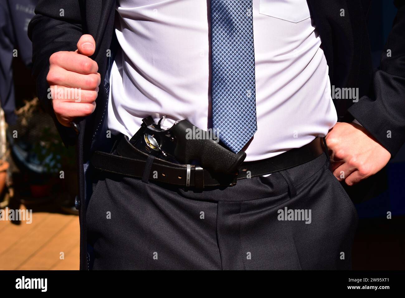 Uomo che indossa un completo vestito nero, una camicia bianca e una cravatta azzurra che mostra il revolver cromato nascosto sotto la cintura Foto Stock