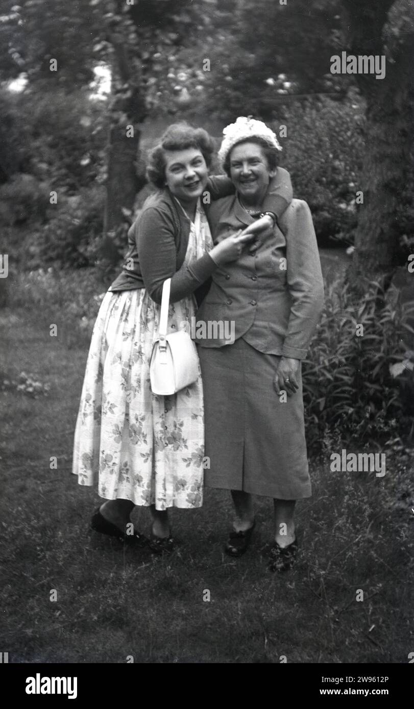Anni '1950, storica, mia mamma....in piedi fuori, una giovane donna che indossa un lungo abito a motivi floreali che dà a sua madre un abbraccio amorevole, Inghilterra, Regno Unito. Foto Stock