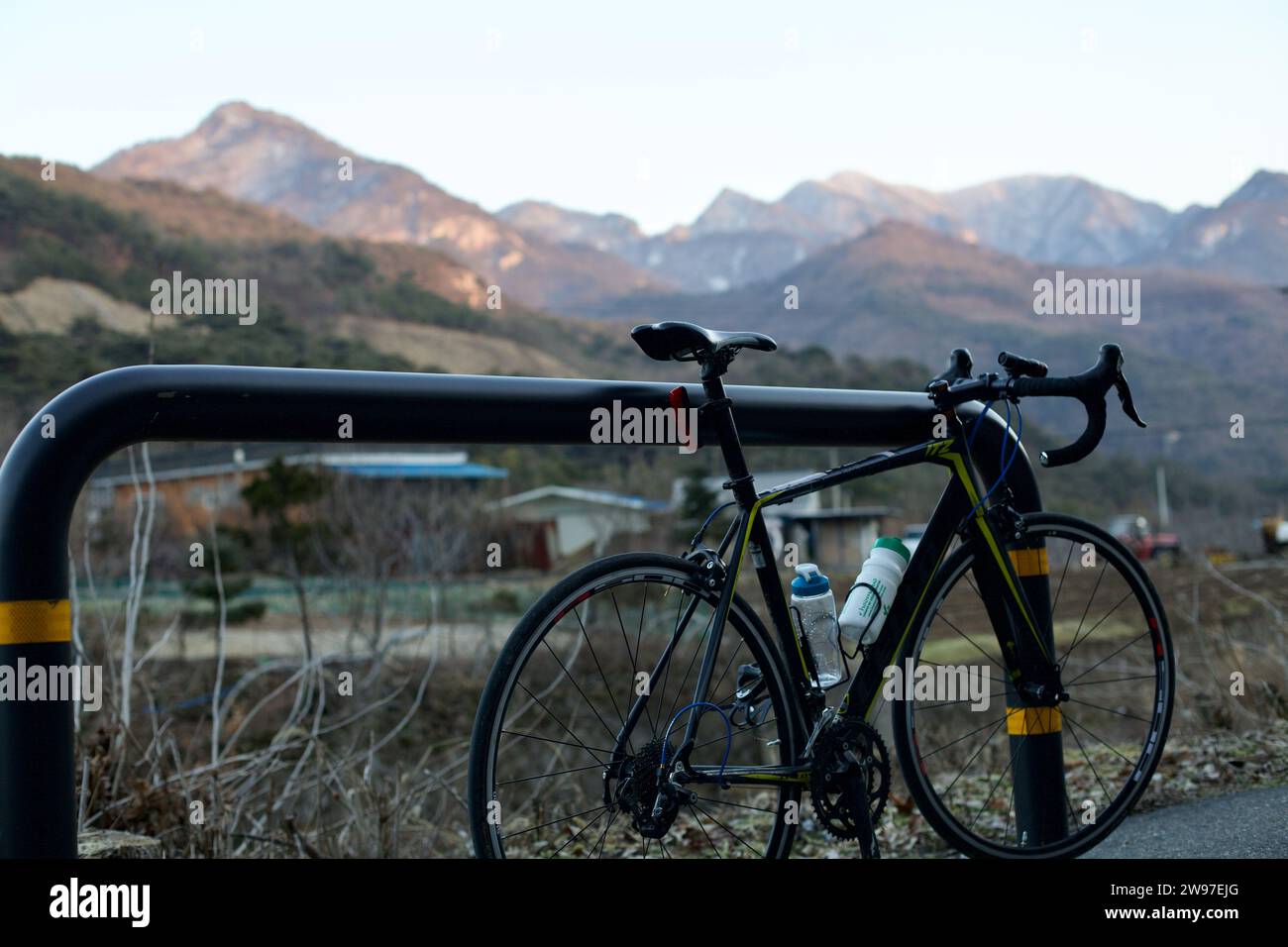 Chungju, Corea del Sud - 22 febbraio 2022: Una bicicletta si appoggia contro un palo di metallo al bordo della strada, con la splendida catena montuosa Sobaek e la sua fattoria Foto Stock