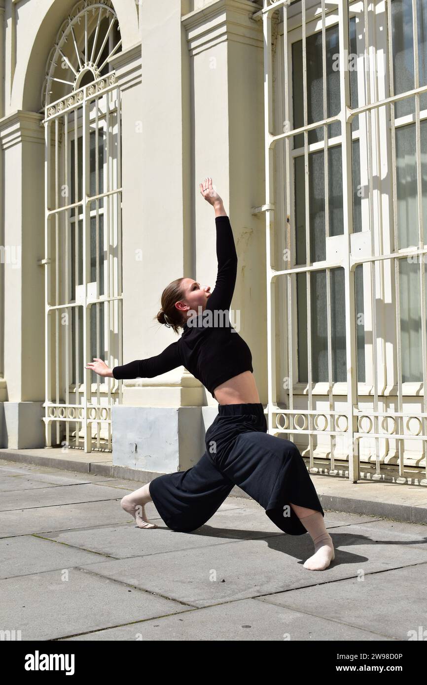 Giovane donna in abito nero che esegue un movimento di danza contemporanea davanti alla finestra con barre bianche Foto Stock