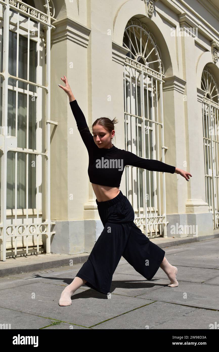 Giovane donna in abito nero che esegue un movimento di danza contemporanea davanti alla finestra con barre bianche Foto Stock