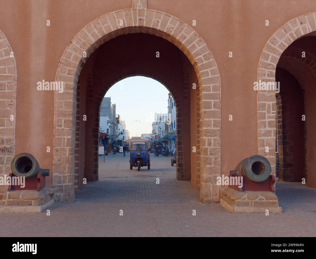 I cannoni accanto alle arcate come veicolo a tre ruote arrivano nella strada principale della medina di Essaouira, in Marocco. 24 dicembre 2023 Foto Stock
