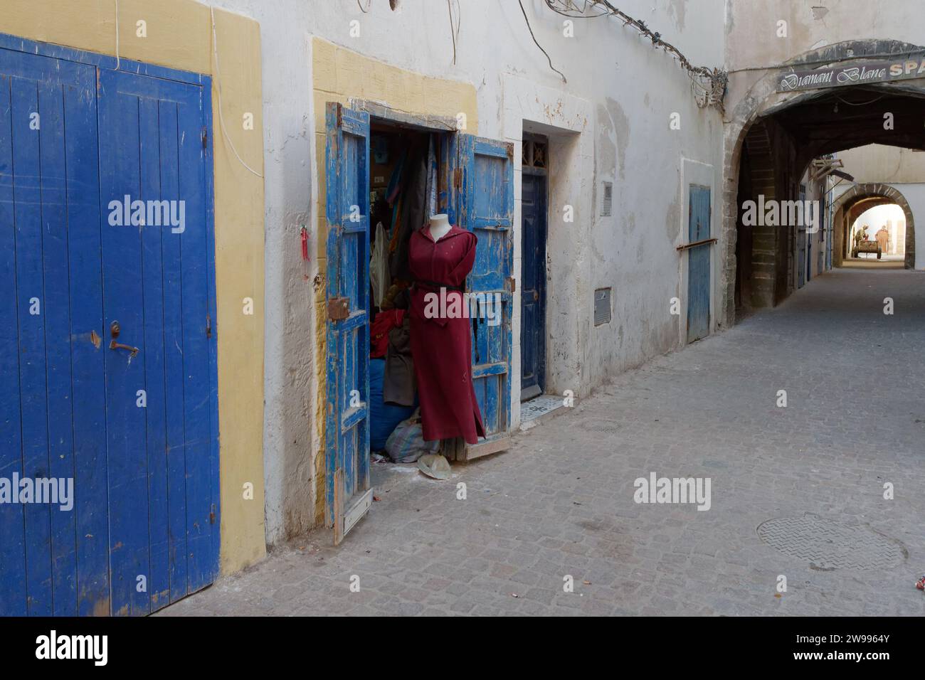 Manichino senza testa con abito rosso in una strada storica nella medina con porte e archi blu a Essaouira, Marocco. 24 dicembre 2023 Foto Stock