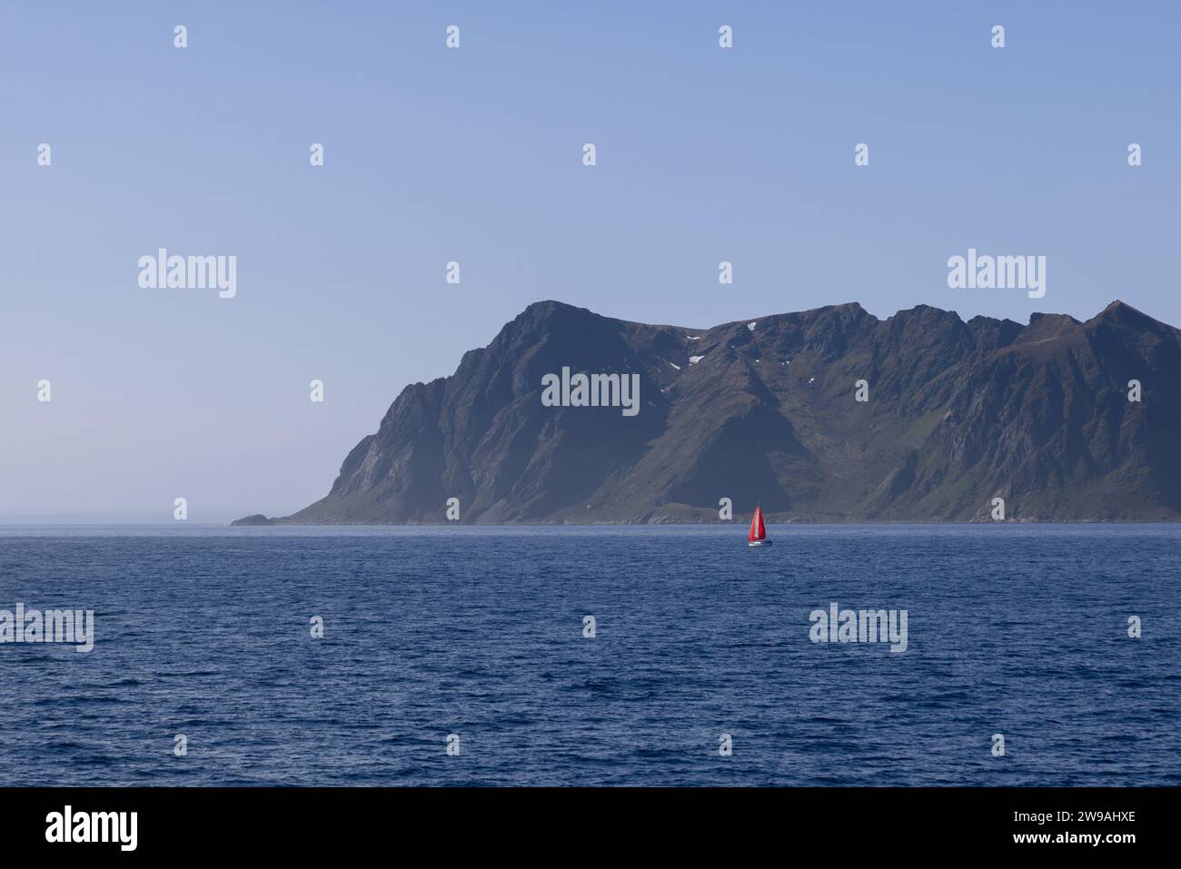 Una vista pittoresca del Mare del Nord e una cresta di montagna che torreggia su acque calme, con una barca a vela che porta una vela rossa vicino all'Isola di Lofoten, sotto un bl Foto Stock