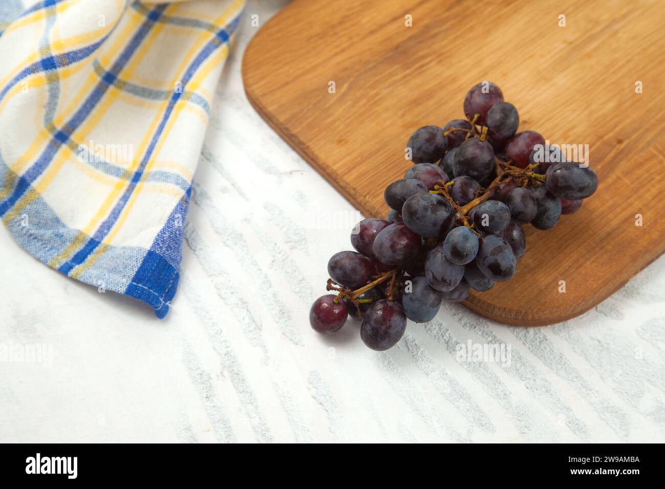 Tagliere con grande mazzo di uva blu scuro maturo su bianco. Uva da vino viola con asciugamano da cucina blu su fondo di legno bianco. Foto Stock