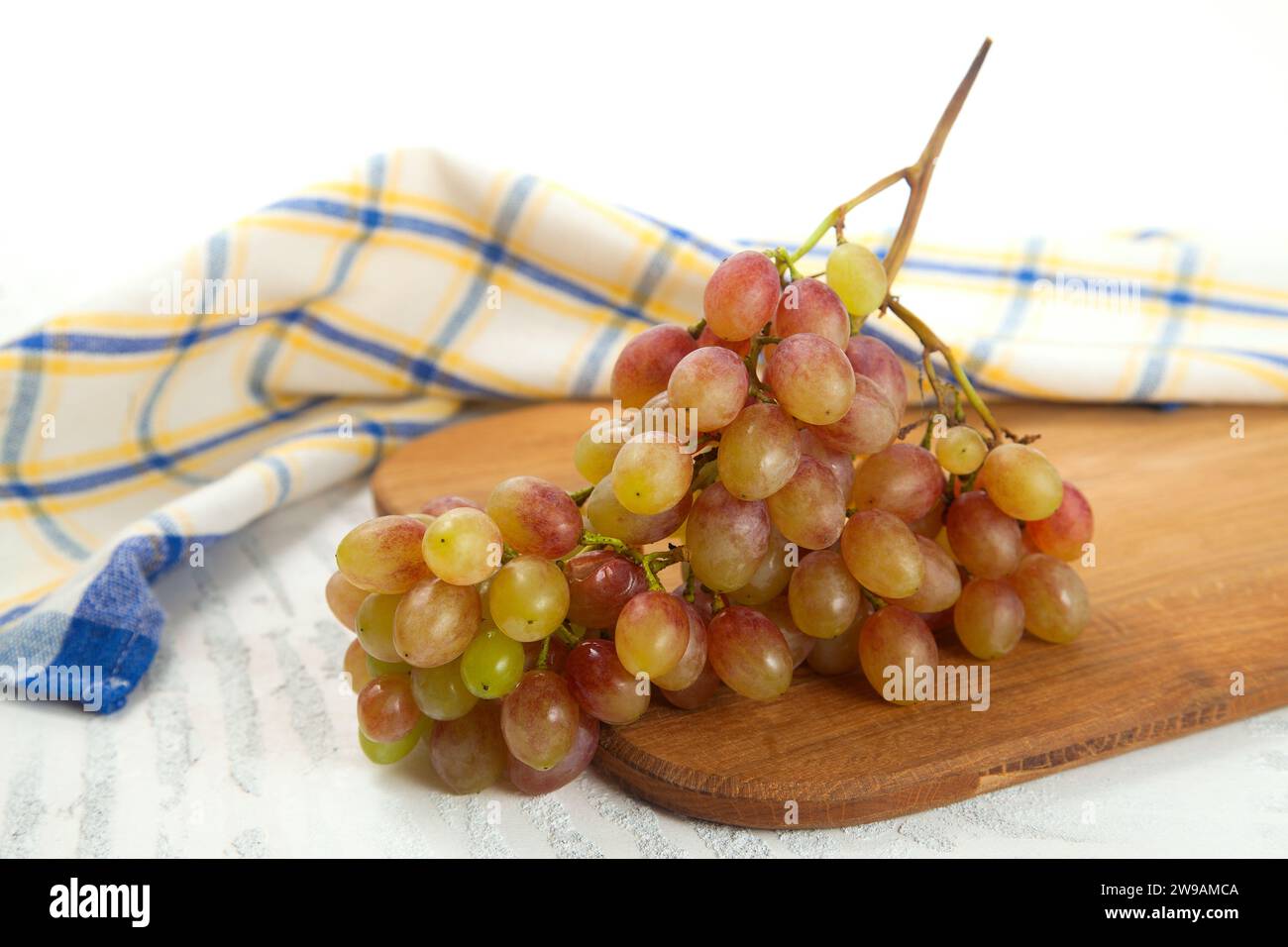 Tagliere con grande mazzo di uva rosa matura su bianco. Uva da vino rosa con asciugamano da cucina blu su fondo di legno bianco. Foto Stock