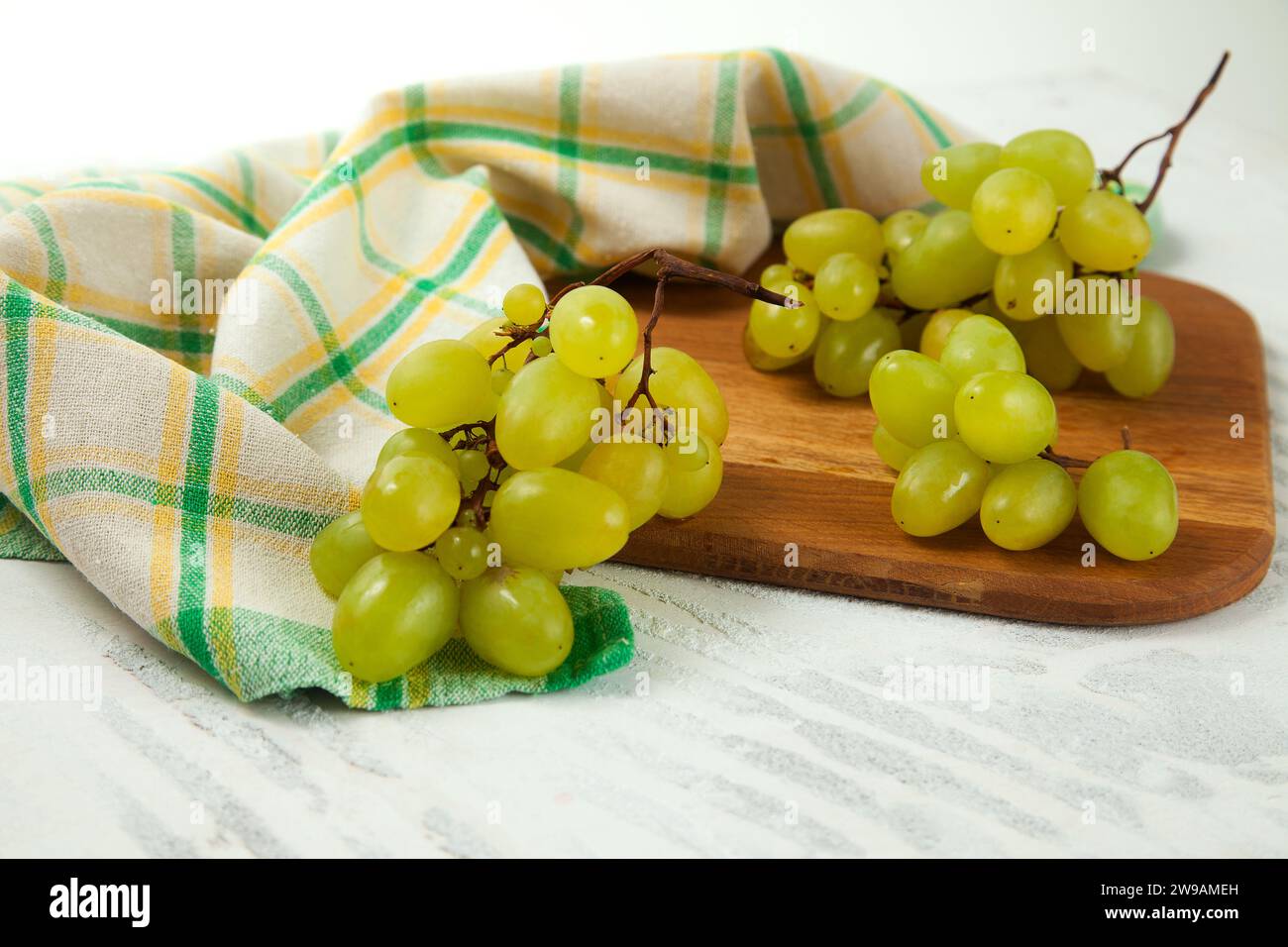 Tagliere con grande mazzo di uva verde matura su bianco. Uva da vino bianco con asciugamano da cucina verde su fondo di legno bianco. Foto Stock