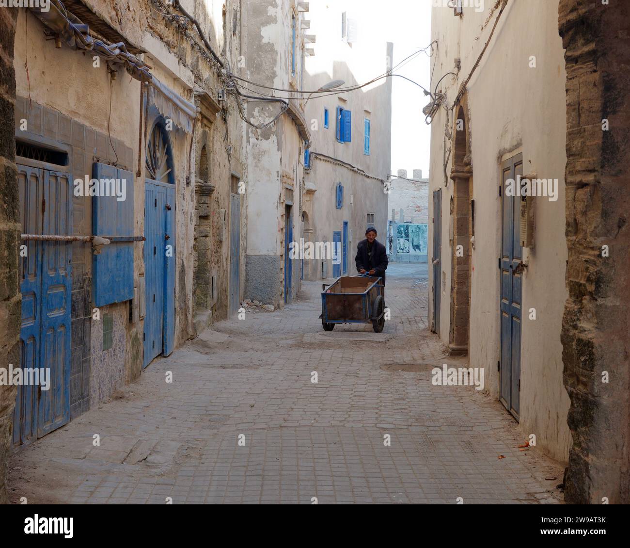 Un anziano spinge un carro di legno lungo una strada polverosa nella medina con porte e persiane di legno blu a Essaouira, in Marocco. 26 dicembre 2023 Foto Stock