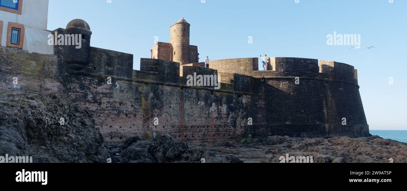 La coppia si trova alle mura e alla fortezza di Essaouira. La donna si fa un selfie mentre il vento soffia la gonna, Marocco. 26 dicembre 2023 Foto Stock