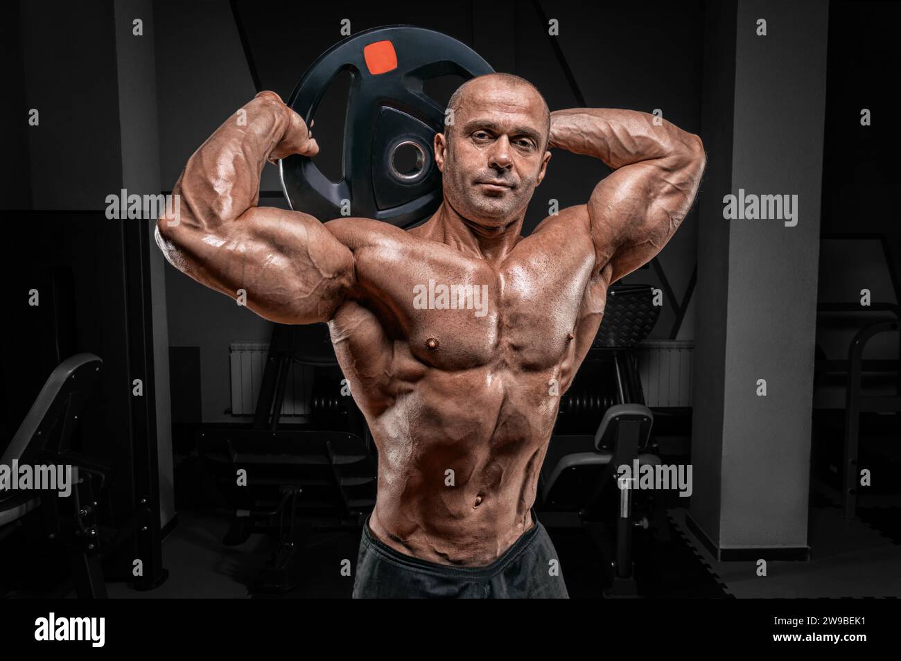 Uomo muscoloso che posa in palestra con un disco a campana. Concetto di allestimento e sollevamento potenziato. Supporti misti Foto Stock