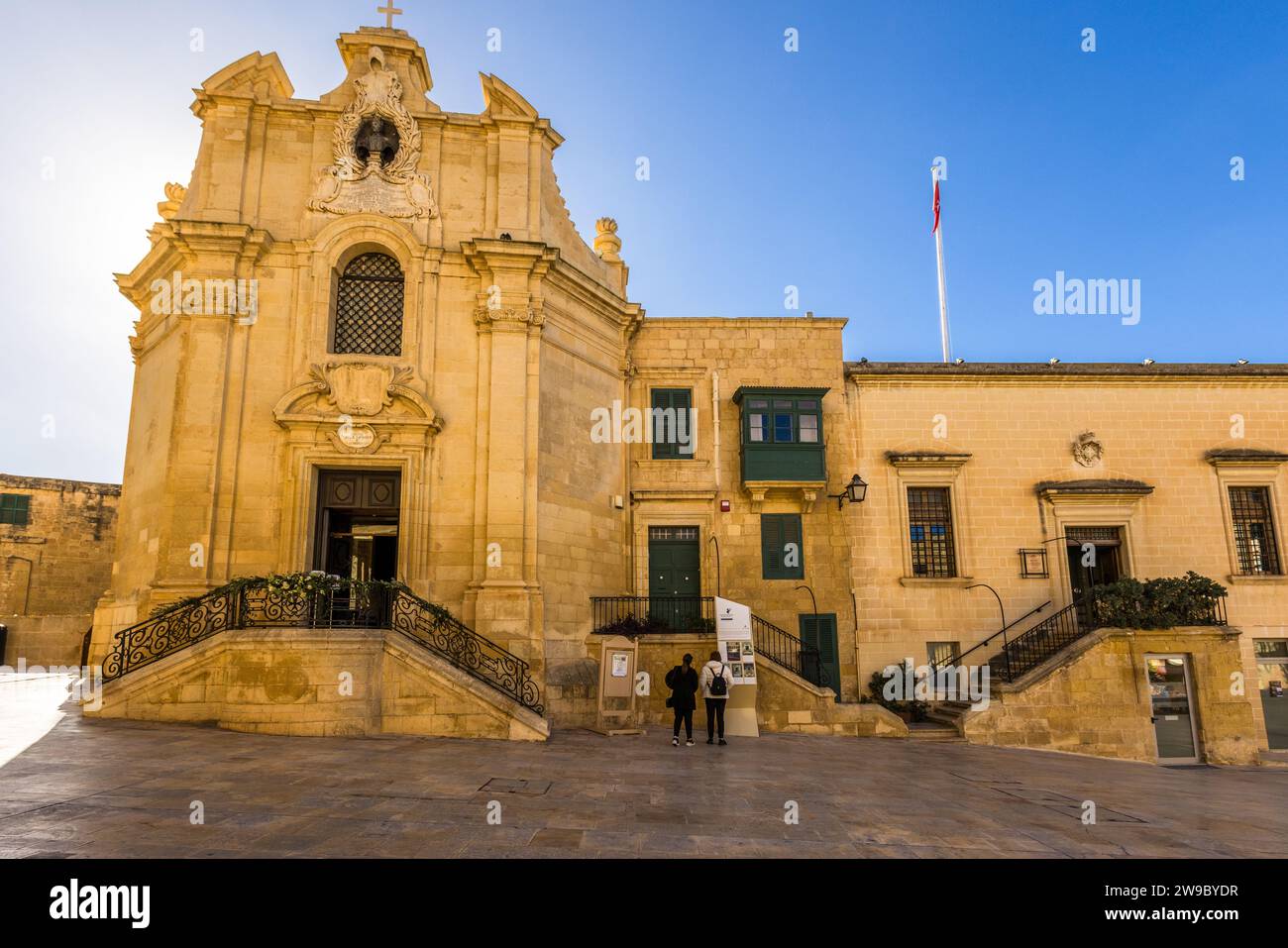 L'Associazione Maltese dell'Ordine di Malta ha sede in questo complesso edilizio su Piazza Jean de Valette a La Valletta, Malta – una facciata di chiesa barocca sormontata da una croce accanto a un edificio più semplice con persiane verdi e la bandiera maltese. Foto Stock