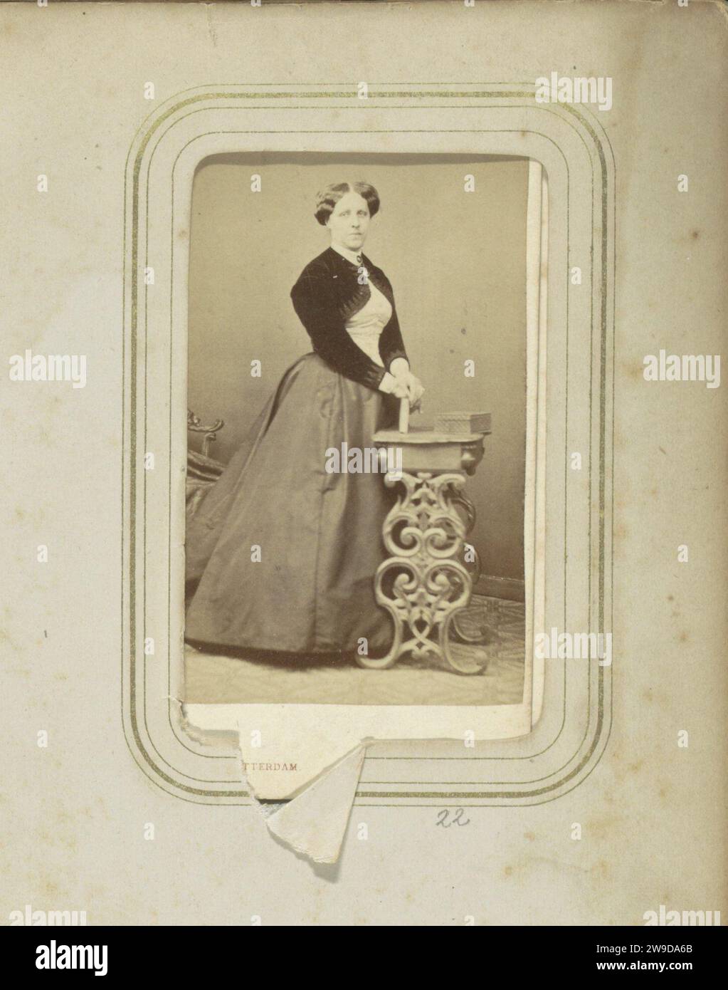 Ritratto di una donna con una gonna lunga e un bolero al tavolo, c. 1860 - c. 1880 scheda visita parte dell'album di famiglia di H. W. F. Ligtenberg e L. M. Swart con 149 foto e cartes-de-visit. Rotterdam cartone. supporto fotografico per la stampa di persone storiche da parte di albumen. famiglia, relazione, discendenza. requisiti studio  fotografo. indumenti per la parte superiore del corpo. indumenti per la parte inferiore del corpo. gonna. Stili di parrucchiere - AA -  donne l'Aia Foto Stock