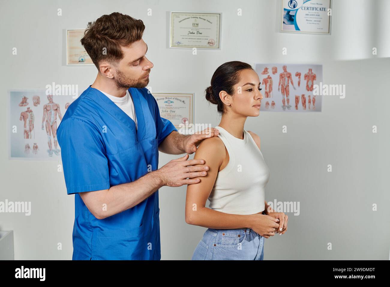 bel medico in costume medico blu che controlla i muscoli della sua giovane paziente femminile, assistenza sanitaria Foto Stock