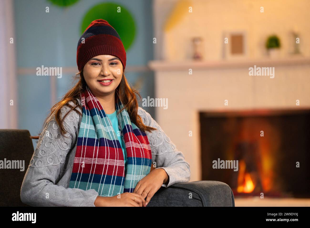 Ritratto di una donna indiana sorridente con un caldo abbigliamento invernale che guarda la macchina fotografica sul divano a casa - concetto di relax, spensieratezza e benessere Foto Stock
