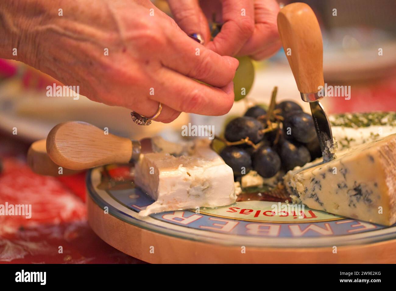 Una donna che prende l'uva dal centro di un piatto di formaggio, con un cuneo stilton con un coltello da formaggio e una fetta di brie cremosa Foto Stock