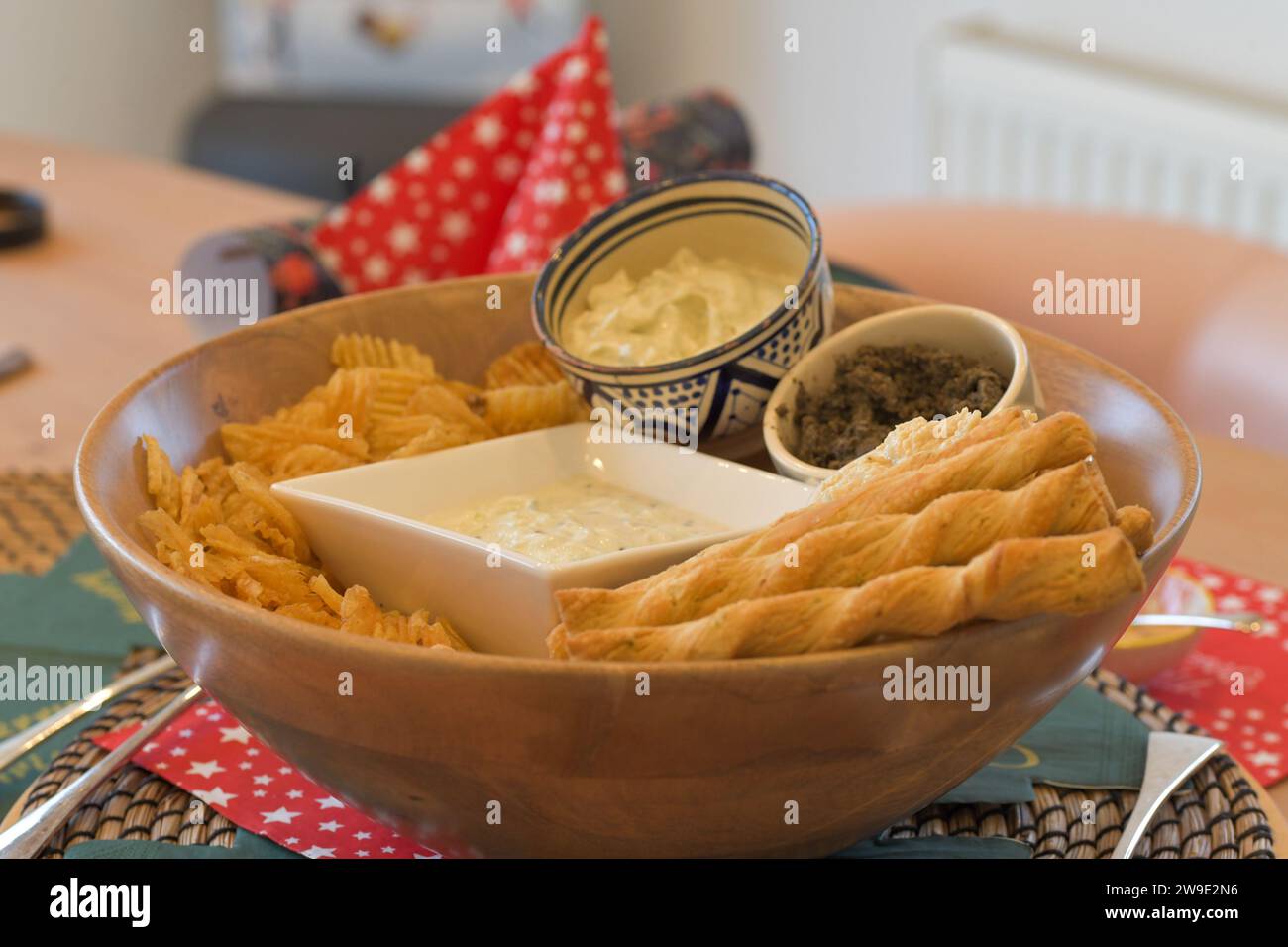 Una ciotola di legno piena di patatine, cannucce e salse al formaggio, con tavoli festivi Foto Stock