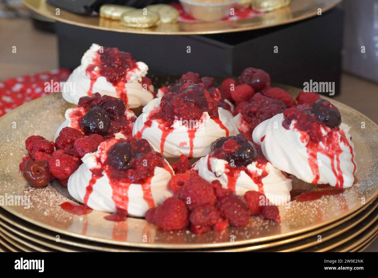 La meringa di lampone e ciliegia nera nidifica su una pila di piatti d'oro, con una spolverata di cristalli di zucchero Foto Stock