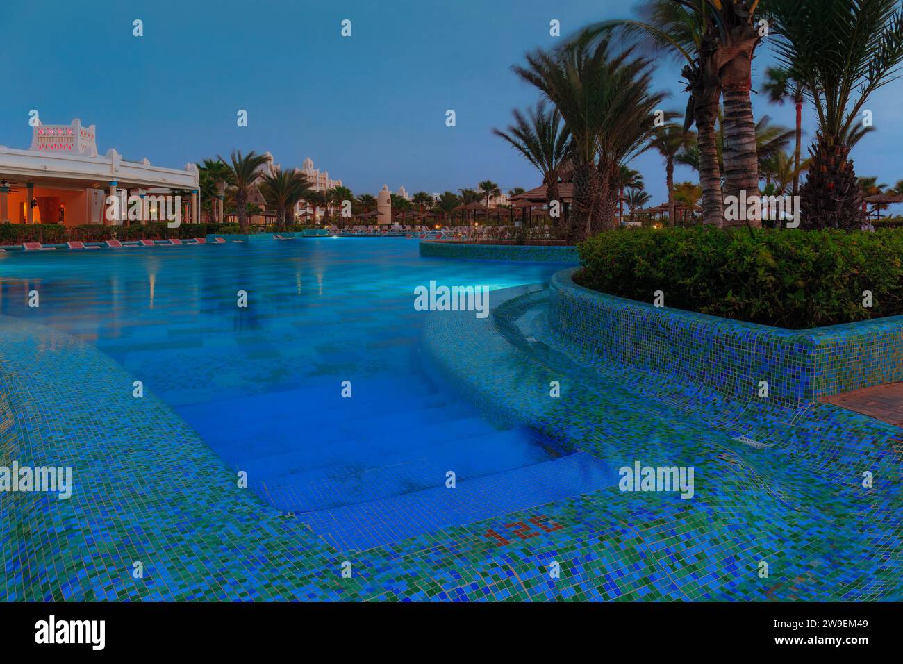 Una piscina dell'hotel si illumina di mosaico blu, incorniciata da sagome di palme e da un vivace ristorante dell'hotel in lontananza. Oasi tropicale notturna. Foto Stock