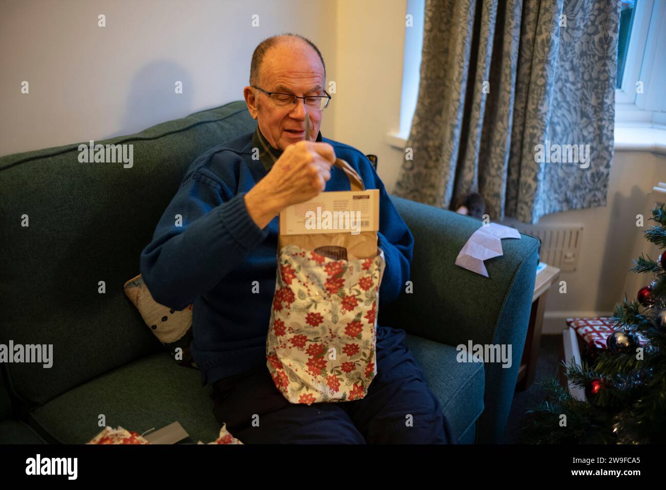 Uomo in pensione di anni settanta che apre un regalo di natale seduto su un divano accanto all'albero di Natale in una casa residenziale, Inghilterra, Regno Unito Foto Stock