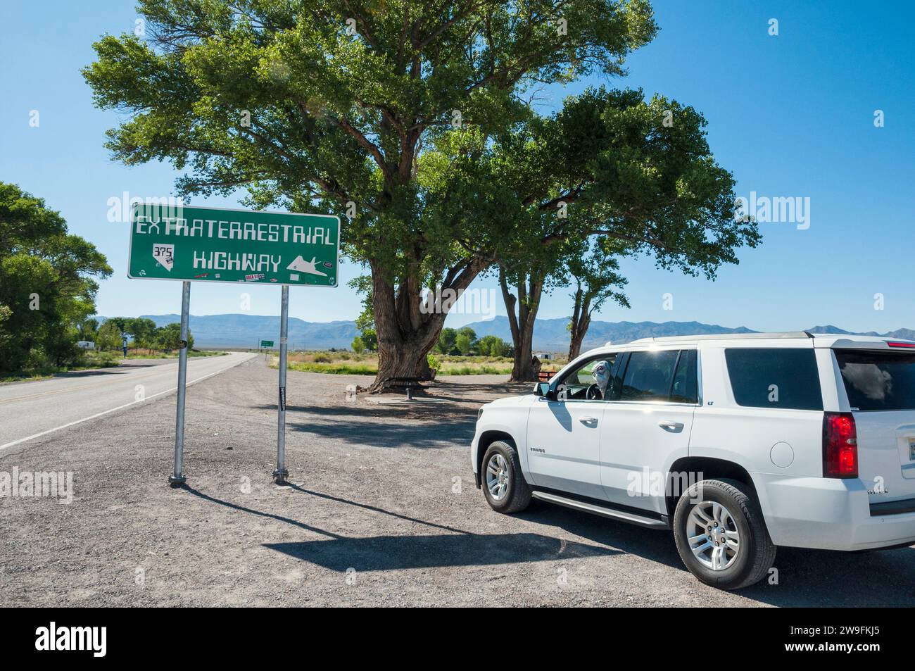 Alieno al volante di una Chevy Tahoe, guardando indietro fuori dal finestrino del sedile del conducente, accanto a un cartello stradale extraterrestre Foto Stock