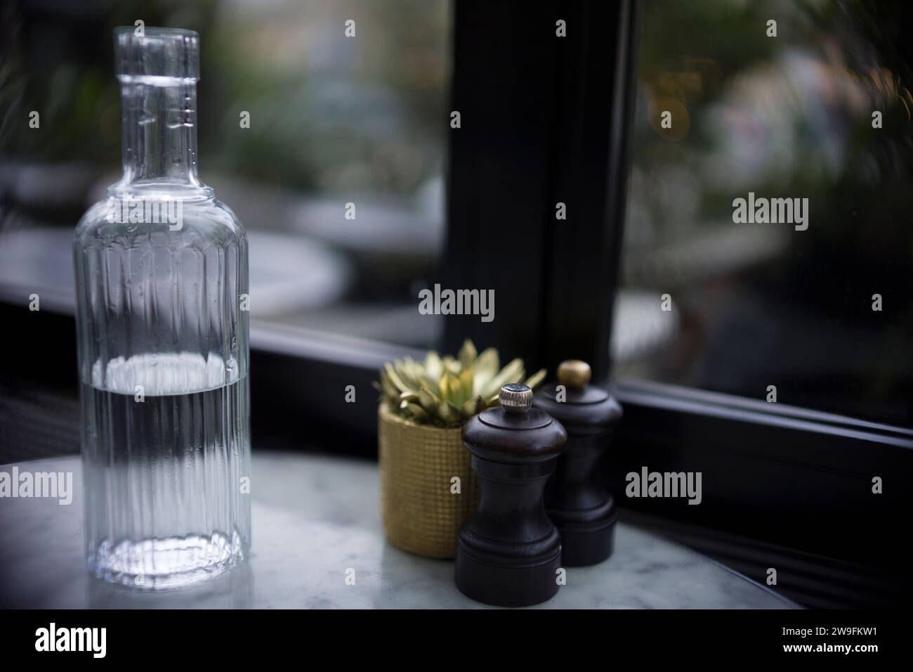 Bottiglia d'acqua in vetro con design a coste, macinacaffè per sale e pepe e una piccola pianta in vaso su un davanzale con sfondo morbido. Foto Stock