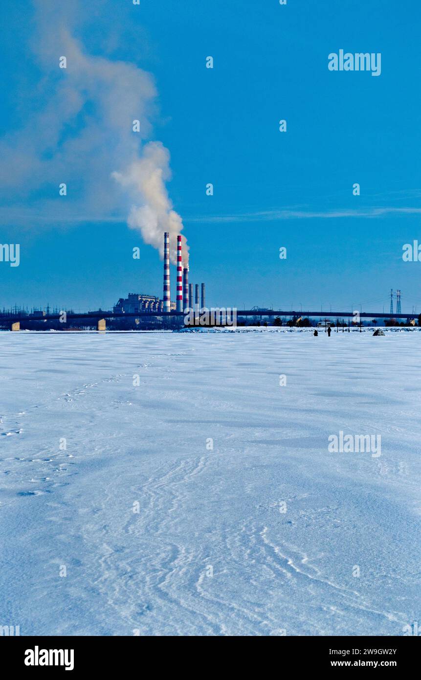 Una vecchia centrale elettrica a carbone che emette fumo tossico nell'atmosfera, paesaggio urbano industriale invernale con fiume ghiacciato Foto Stock