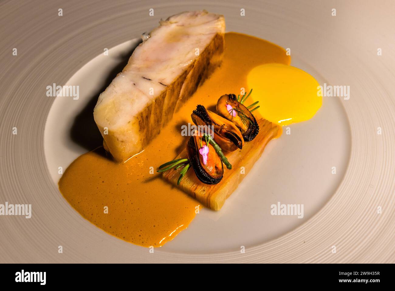 Un pezzo di pesce bianco è impiattato nel ristorante stellato Michelin Noni a Valletta, Malta, insieme a cozze grigliate su porri, circondato da una salsa arancione e una cucchiaiata gialla. Lo chef Jonathan Brincat gestisce il ristorante di Republic Street dalla sua apertura nel 2017; nel 2020 è diventato uno dei primi tre ristoranti di Malta a ottenere una stella Michelin. Foto Stock