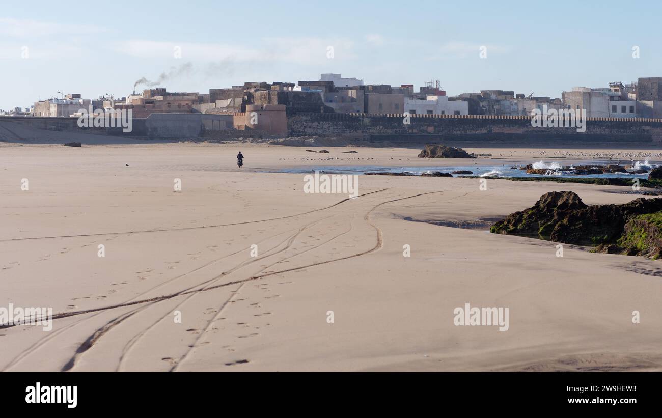 Qualcuno va in moto su una spiaggia sabbiosa e si dirige verso la Medina e le mura cittadine di Essaouira, in Marocco. 28 dicembre 2023 Foto Stock