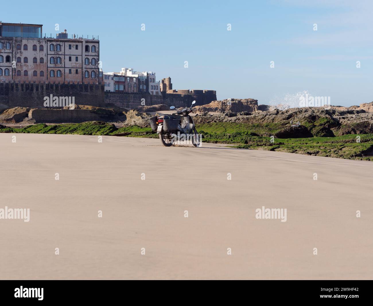 IMotorbike parcheggiata su una spiaggia sabbiosa con formazioni rocciose e la storica Medina alle spalle nella città di Essaouira, Marocco. 28 dicembre 2023 Foto Stock
