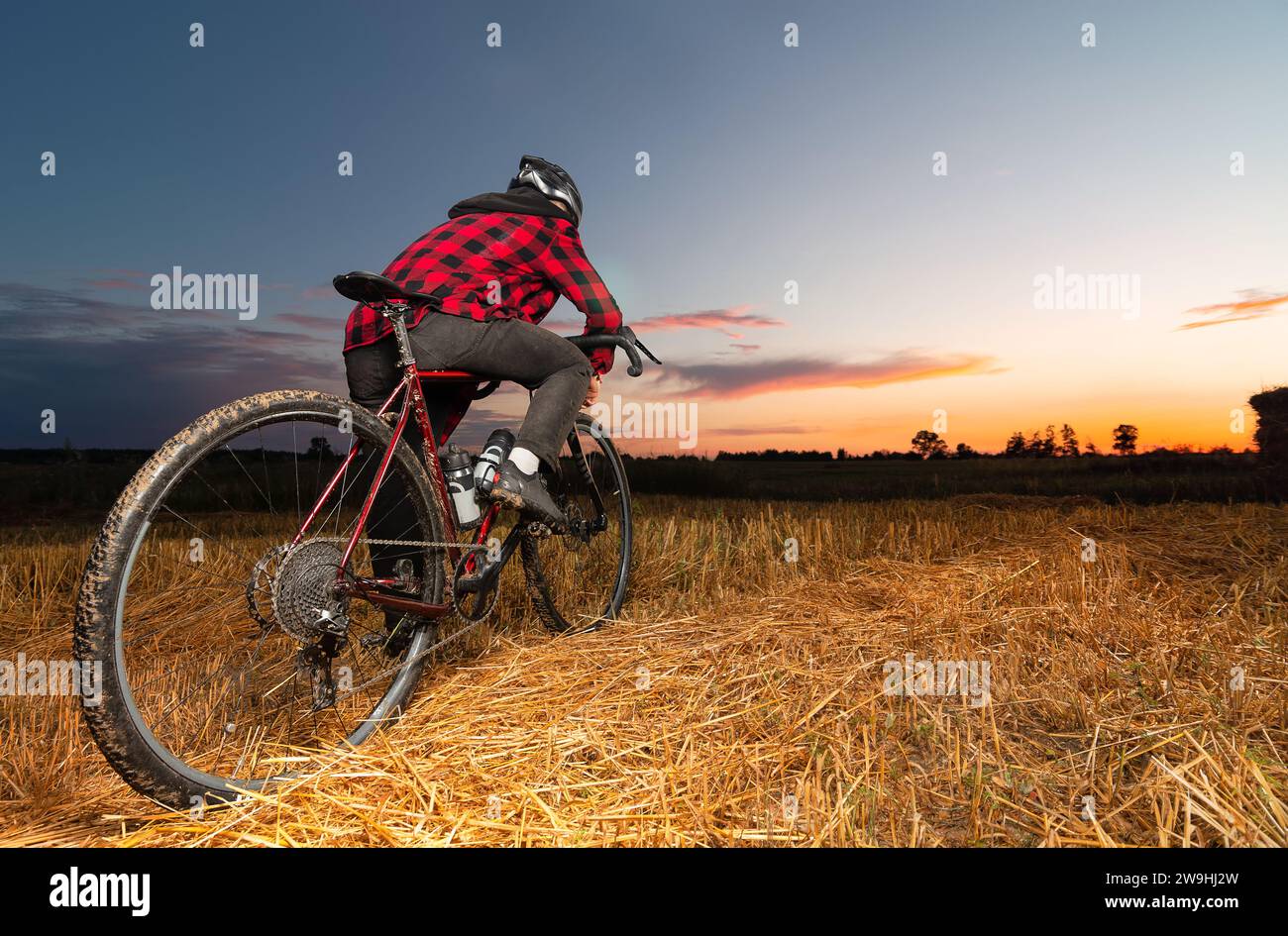 Un ciclista su una bici di ghiaia si trova in un campo sullo sfondo del tramonto. Vista angolare bassa. Foto Stock
