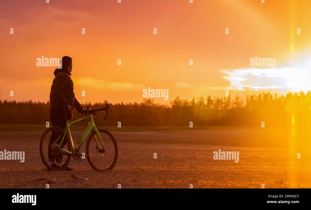 L'uomo con una bici in ghiaia si trova in un campo su uno sfondo splendido al tramonto. Splendido paesaggio con mountain bike. Foto Stock