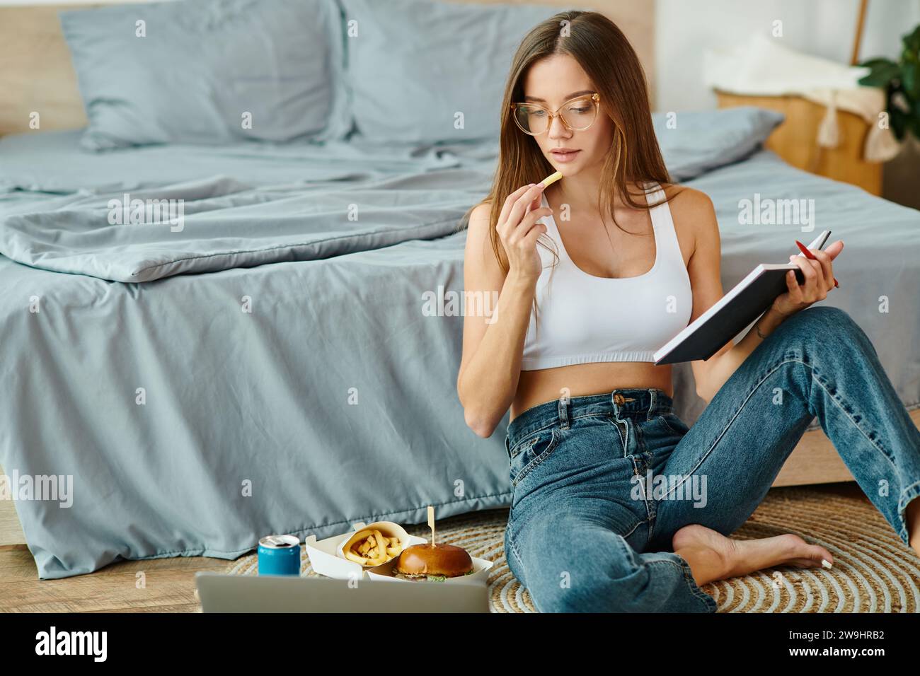 giovane donna premurosa con un abito informale seduta sul pavimento mentre lavora a distanza e mangia patatine fritte Foto Stock