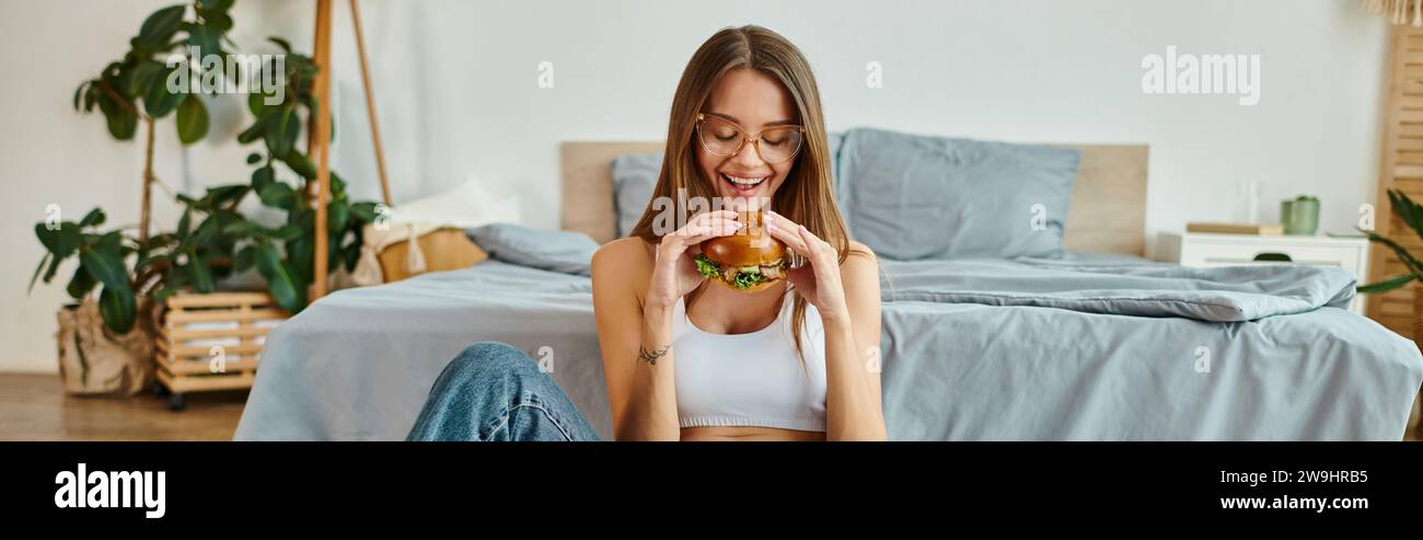 donna allegra e attraente con occhiali e capelli lunghi che si gusta un delizioso hamburger mentre è a casa, striscione Foto Stock