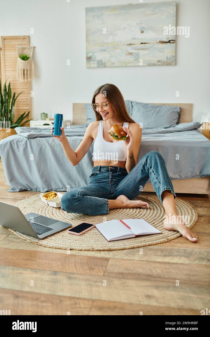 donna felice e attraente, con abbigliamento casual, seduta a terra e mangiando hamburger mentre lavora sul suo computer portatile Foto Stock